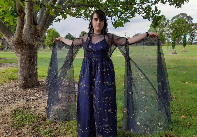 Image of Ella in her formal dress. It is a dark blue, with embroidered stars and constellations, she has her arms outstretched to show the length of the sleeves.